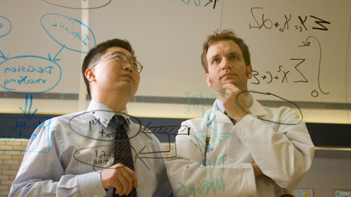 Two men study a diagram they've drawn with marker on a glass panel